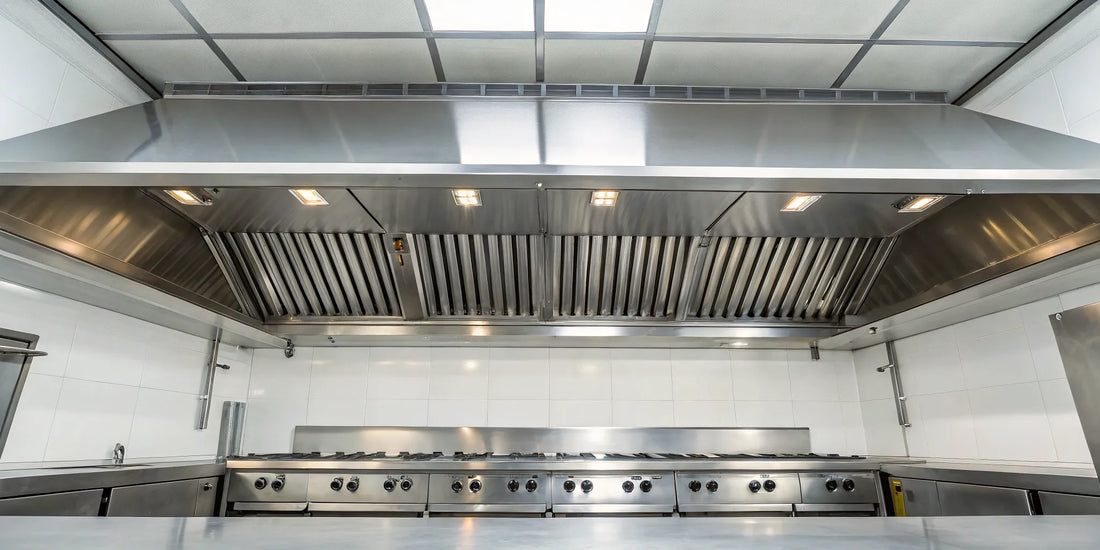 A stainless steel grease duct fan and exhaust hood over a commercial kitchen cooking line.